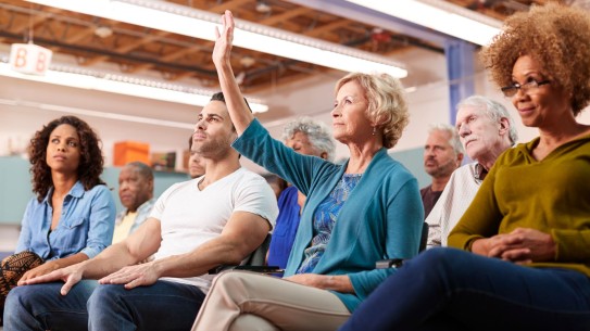 People sitting in a meeting, one with arm raised for a question