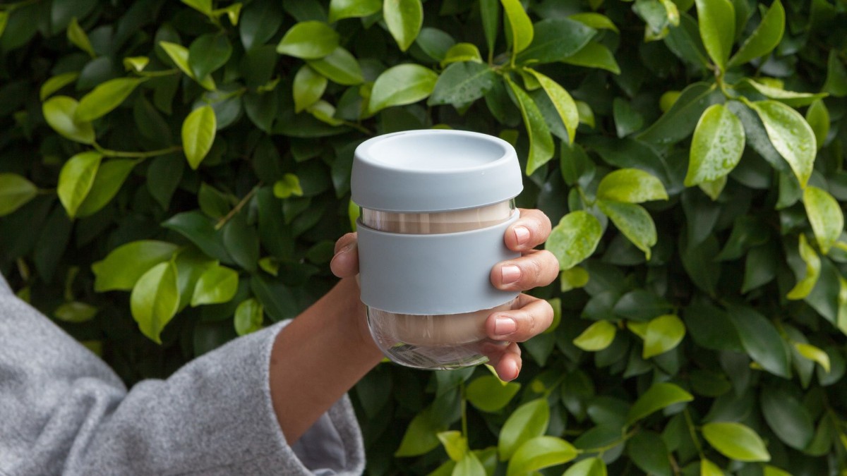 photo of a hand holding a glass reusable coffee cup in front of a leafy plant