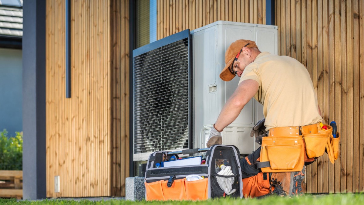 A man in a toolbelt inspecting a heat pump unit on the outside of a wooden clad house