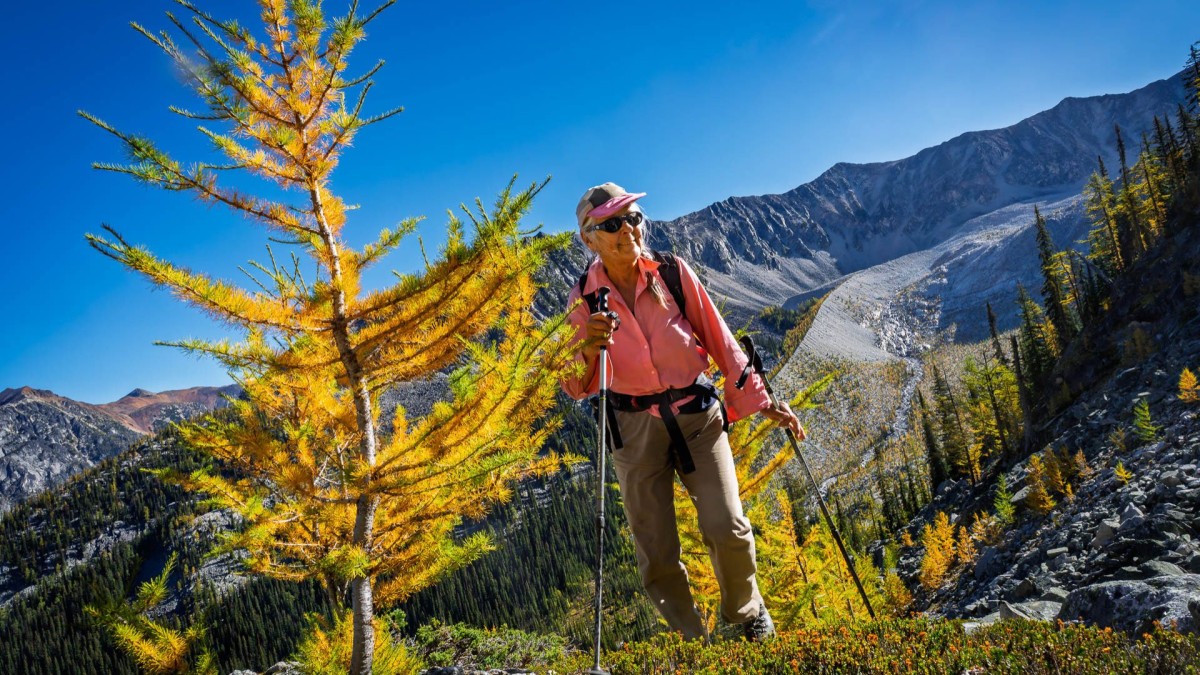 Photo of Baiba dressed in a pink shirt and hat, hiking among mountains and golden larches