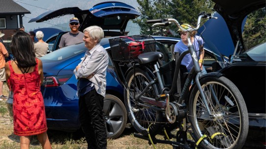 electric cars with doors open, one with an electric bike loaded on the bike rack, people looking at them and talking