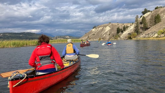 view down river with two people in a red canoe in foreground and other canoes and kayaks further ahead