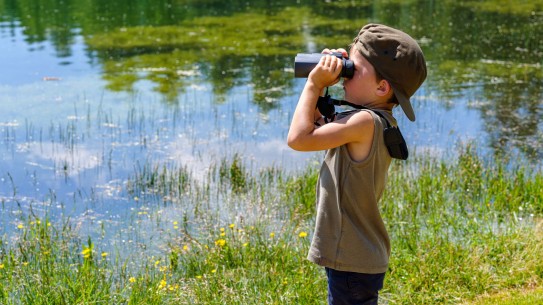 young boy looking through binoculars standing at the edge of a pond