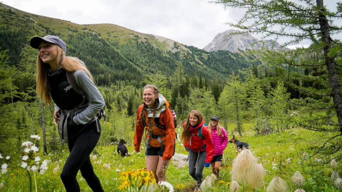 four young women in bright jackets hiking through mountain meadow with two dogs playing