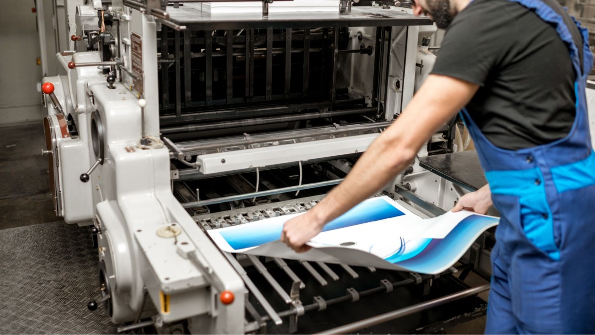 a man in blue overalls lifting a large sheet of paper out of a printing machine