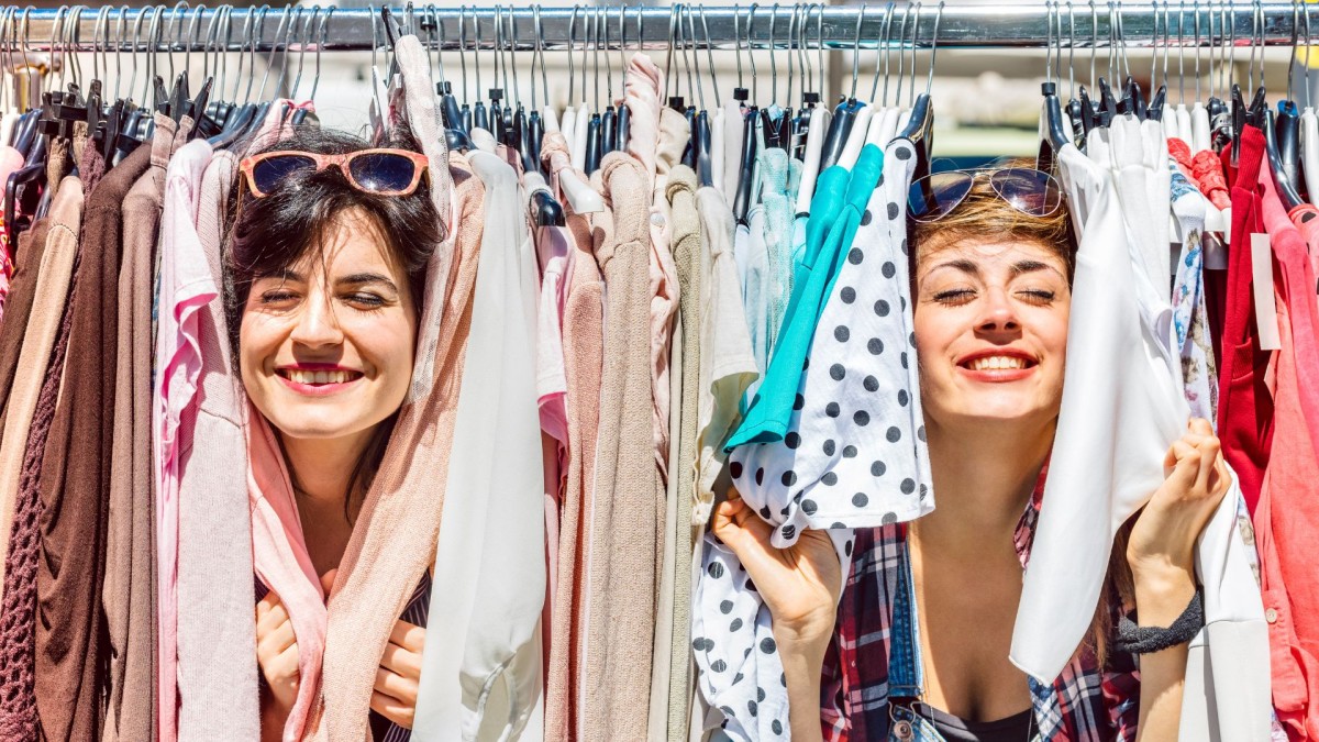 two smiling ladies peeking through a rack of colourful clothes